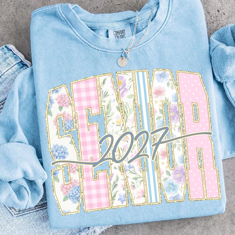 May include: A light blue sweatshirt with the word "SENIOR" in large, gold-outlined letters. The letters are filled with floral, gingham, and striped patterns. The year "2027" is written below in a gray script. A silver necklace with a small pendant is visible.