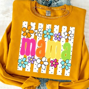 May include: Mustard yellow sweatshirt with a graphic that says "mama" in colorful bubble letters. The word is set against a white background with vertical stripes and dotted accents, surrounded by colorful flower illustrations.