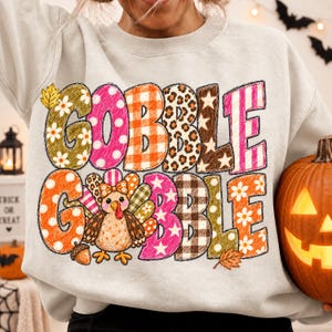 May include: A light gray crewneck sweatshirt featuring the text GOBBLE GOBBLE in large, multi-patterned letters. The design includes a cartoon turkey, a small acorn, and a fallen leaf, with each letter filled with various prints like plaid, leopard, and floral.