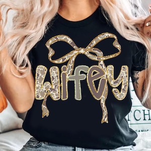 Könnte beinhalten: Schwarzes T-Shirt mit dem Wort "Wifey" in einem Camouflage-Muster mit goldenen Verzierungen. Ein passendes Schleifendesign befindet sich über dem Wort. Das Shirt ist ein lässiger Stil.