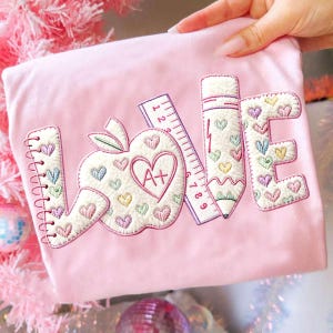 May include: Pink t-shirt with the word "LOVE" embroidered in white, featuring an apple, pencil, and ruler design. The apple has a heart with "A+" inside. The letters and apple are decorated with colorful hearts.