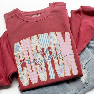 May include: A maroon sweatshirt with the word "MAMA" in large, colorful, patterned letters. The letters are outlined in gold and filled with floral, gingham, and polka dot designs. The word "custom" is written in cursive below.
