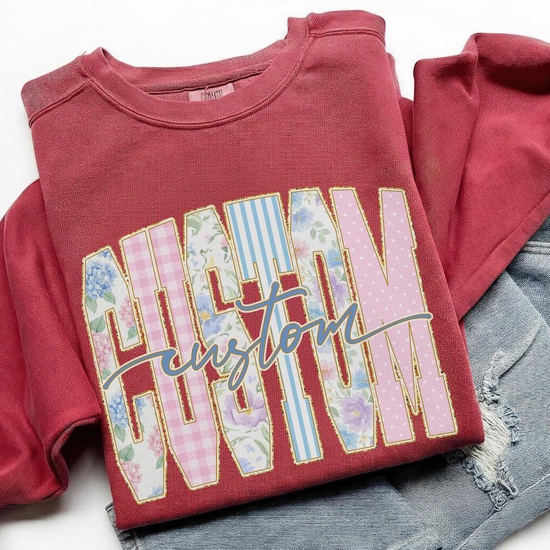 May include: A maroon sweatshirt with the word "MAMA" in large, colorful, patterned letters. The letters are outlined in gold and filled with floral, gingham, and polka dot designs. The word "custom" is written in cursive below.