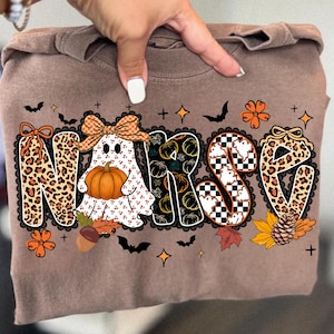 May include: Brown long-sleeve shirt with the word "NURSE" in a Halloween-themed design. The letters are decorated with leopard print, pumpkins, and a ghost holding a pumpkin. Black bats, stars, and fall leaves complete the design.