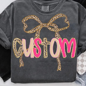 Puede incluir: Sudadera gris oscuro con un lazo con estampado de leopardo y la palabra "CUSTOM" en letras rosas y beige. La sudadera tiene cuello redondo y mangas largas. El lazo está delineado en dorado.