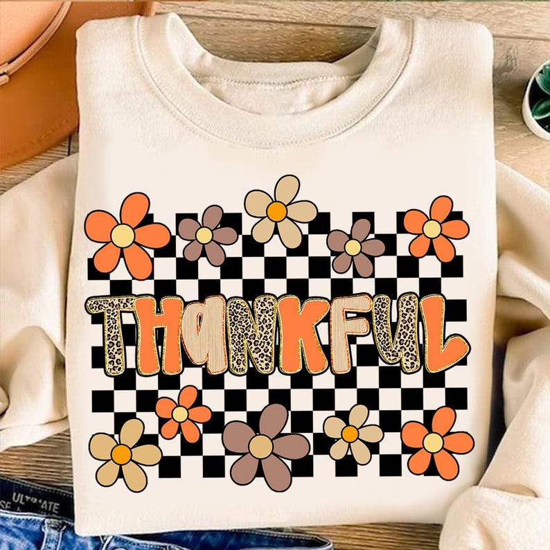 May include: Cream-colored sweatshirt with the word "THANKFUL" in orange and leopard print lettering. The design includes orange, brown, and tan flowers, and a black and white checkered pattern.