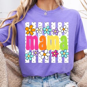 May include: A lilac-colored t-shirt with the word "mama" in large, colorful letters. The word is surrounded by a pattern of white vertical stripes, black dots, and colorful flowers. The shirt has short sleeves.
