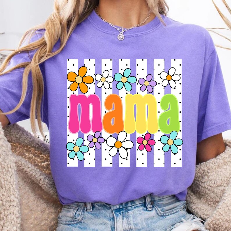 May include: A lilac-colored t-shirt with the word "mama" in large, colorful letters. The word is surrounded by a pattern of white vertical stripes, black dots, and colorful flowers. The shirt has short sleeves.
