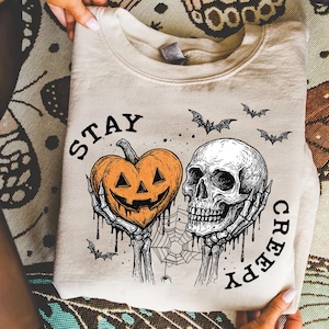 May include: Beige sweatshirt with a Halloween-themed graphic. The design features a jack-o'-lantern heart and a skull held by skeletal hands, with the words "STAY CREEPY" and bats. A spooky and festive design.