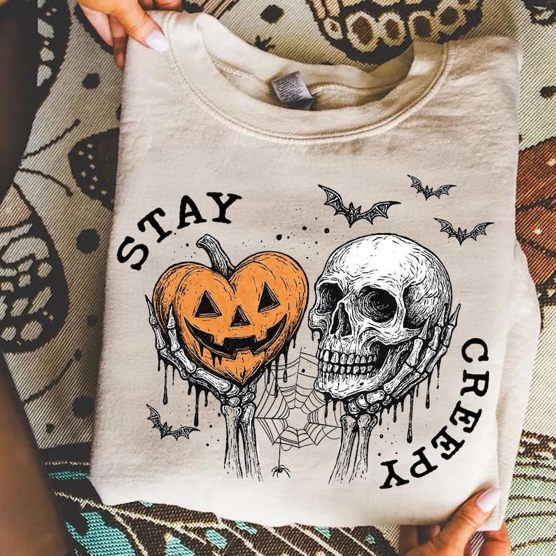 May include: Beige sweatshirt with a Halloween-themed graphic. The design features a jack-o'-lantern heart and a skull held by skeletal hands, with the words "STAY CREEPY" and bats. A spooky and festive design.
