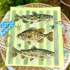 May include: A canvas print featuring three detailed illustrations of fish: a catfish, a bass, and a trout. The background is a bright yellow-green with vertical stripes. Decorative stars and jewelry are placed around the canvas.