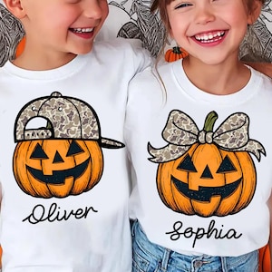 May include: Two white t-shirts with Halloween-themed pumpkin designs. One shirt features a pumpkin wearing a camouflage baseball cap, the other a pumpkin with a camouflage bow. The names "Oliver" and "Sophia" are written below the pumpkins.