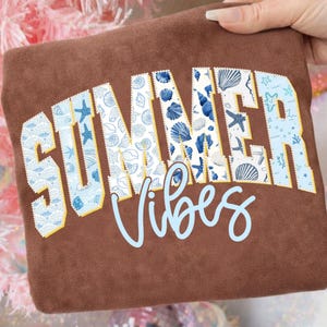 Puede incluir: Una sudadera marrón con las palabras "SUMMER Vibes" en azul y blanco con diseño de conchas marinas. Las letras tienen un borde amarillo. La sudadera es sostenida por una mano.