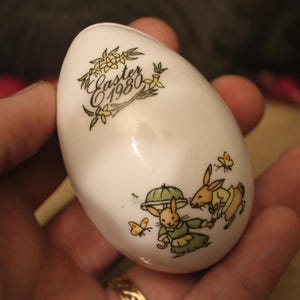 Puede incluir: Huevo de Pascua de cerámica blanca con un diseño decorativo con conejitos, mariposas y el texto "Easter 1980". El huevo se sostiene en una mano y tiene un acabado brillante. Se ve un anillo dorado en la mano.