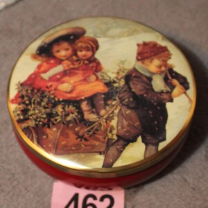 May include: A round, red trinket box with a gold rim and a lid featuring a vintage illustration of children in winter clothing. The lid's artwork depicts two children in red dresses and a boy carrying a bundle of sticks. A pink tag with the number 462 is visible.