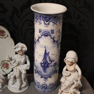 Może przedstawiać: Kolekcja zabytkowych przedmiotów porcelanowych, w tym wysoki niebiesko-biały wazon z motywem żaglówki, dekoracyjny talerz z parą i figurki. Wazon ma około 25 cm wysokości.