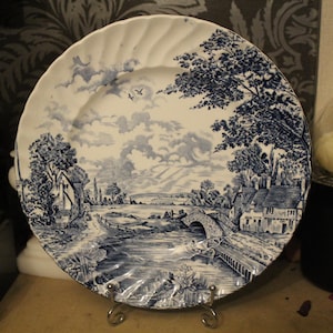 Può includere: Un piatto rotondo in ceramica con un motivo blu e bianco che raffigura un paesaggio rurale. La scena mostra un ponte in pietra su un fiume, casolari, alberi e uccelli nel cielo. Il piatto presenta un bordo ondulato e smerlato.