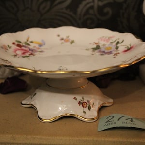 Peut inclure: Un présentoir à gâteau en porcelaine blanche avec un bord festonné et une bordure dorée. Le présentoir présente un motif floral avec des fleurs roses, violettes et jaunes. La base a un motif floral assorti. Diamètre d'environ 20 cm.