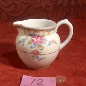 Könnte beinhalten: Cremefarbener Keramik-Milchkännchen mit einem Blumendesign aus rosa Rosen und blauen Vergissmeinnicht. Das Milchkännchen hat einen gebogenen Griff und eine dekorative gold-blaue Bordüre am Rand. Unten befindet sich ein rosa Etikett mit der Nummer 72.