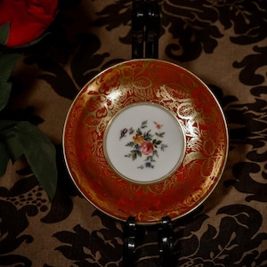 May include: An ornate, antique saucer in red and gold. The saucer has a central white section with a floral design. The red outer section is decorated with gold patterns. A vintage piece, ideal for display or serving tea or coffee.