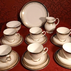 Peut inclure: Service à thé blanc avec bordures dorées et vertes, comprenant tasses, soucoupes, un plat de service, une crémière et un sucrier. L'ensemble est disposé sur une surface rouge, mettant en valeur un design classique.