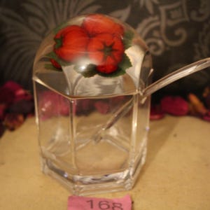 Può includere: Una zuccheriera in vetro trasparente e sfaccettato con coperchio trasparente. Il coperchio presenta una disposizione decorativa di pomodori rossi. Un cucchiaio trasparente è all'interno.