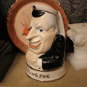 May include: A ceramic character jug shaped like a man wearing a black jacket and a white captain's hat. The figure has a pale complexion, blue eyes, and a wide grin. The base of the jug features the text "DARLING JUG" in black capital letters.