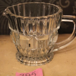 Puede incluir: Una jarra de cristal transparente y facetado con asa y boquilla. La jarra tiene un diseño geométrico decorativo y se asienta sobre una base redonda. El asa es angular y transparente.