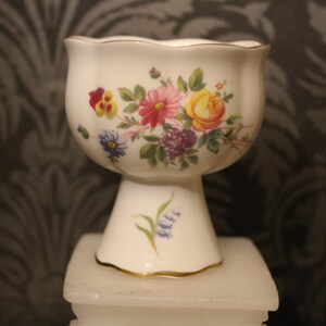 May include: A white porcelain pedestal bowl with a scalloped rim, decorated with a colourful floral design featuring pink, yellow, and purple flowers. The base has a gold trim and a painted bluebell detail.