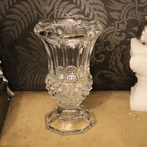 May include: A clear glass vase with a flared top and a diamond-patterned base. The vase has a hexagonal base and vertical ribbed detailing. The background features a dark, patterned wallpaper. The vase is likely around 15 cm tall.
