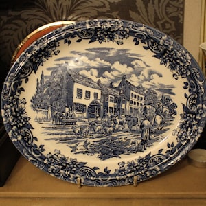 May include: An oval ceramic platter featuring a blue and white transferware pattern. The central scene depicts a rural village with buildings, a flock of sheep, and figures. The wide border is decorated with intricate floral and scroll motifs.