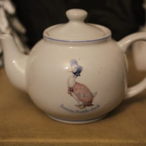 May include: A white ceramic teapot with a lid and handle. The teapot has a blue trim and a duck illustration wearing a bonnet and dress. The text "Jemima Puddle-duck" is below the duck.
