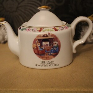 May include: White ceramic teapot with a lid and handle, featuring a round illustration of rabbits by a fireplace. The teapot has a gold-coloured finial and trim, with the text "THE TALES OF HONEYSUCKLE HILL".