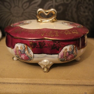 Puede incluir: Una caja de baratijas decorativa de porcelana con tapa. La caja presenta una combinación de colores burdeos y crema con detalles dorados. La tapa tiene un asa en forma de corazón. Los lados de la caja tienen imágenes ovaladas de personas con ropa de época.