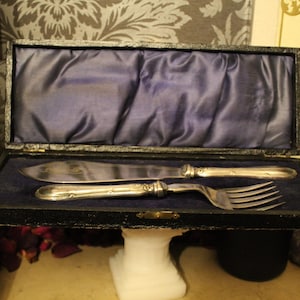 May include: A vintage silverware set, including a knife and fork, presented in a black box with a blue satin lining. The silverware has ornate handles and is displayed on a white pedestal.