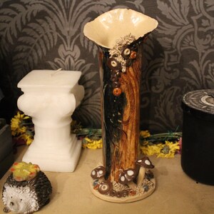May include: A tall, cylindrical ceramic vase with a flared, cream-coloured rim. The body features a textured, wood-like brown and black finish, adorned with small sculpted mushrooms and moss-like details at the base and near the top opening.