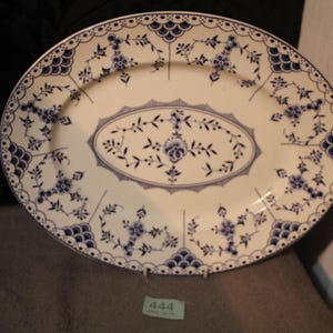 May include: An oval ceramic serving platter featuring a cream-colored base with a detailed blue floral pattern. The rim is decorated with scalloped blue motifs and delicate vines. A small green tag with the number 444 is visible at the bottom.