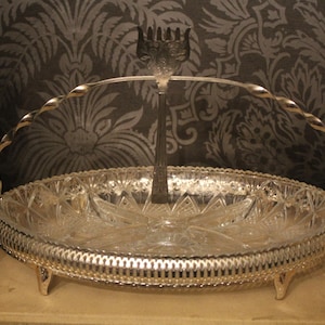 May include: A silver-toned serving dish with a clear, starburst-patterned glass bowl. It features a decorative handle and a central serving utensil. The dish is supported by small feet, adding to its elegant design.