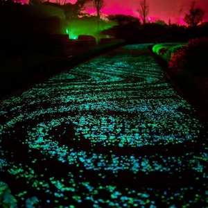 Puede incluir: Un camino bordeado de guijarros verdes y azules brillantes, creando un efecto mágico y luminoso. El camino está rodeado de follaje oscuro y árboles, con un tenue resplandor rosado en el fondo.