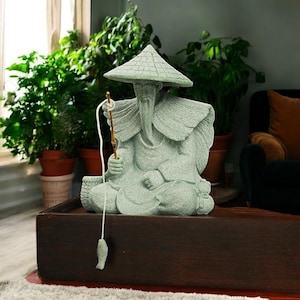Può includere: Statua verde chiaro di un pescatore asiatico seduto, con un cappello conico e una canna da pesca. La statua ha una lunga barba ed è seduta su una scatola di legno. Un piccolo amo a forma di pesce è appeso.