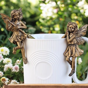 Puede incluir: Dos figuras de hadas color bronce adornan una taza blanca. Una hada sostiene un báculo, la otra apoya la barbilla en sus manos. Las hadas tienen alas y vestidos detallados. La taza tiene un diseño simple y elegante, para un toque caprichoso.