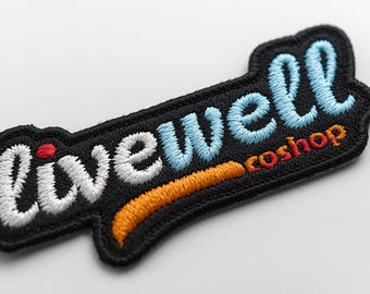 May include: Embroidered patch featuring the text "livewell coshop". The words are stitched in white, light blue, orange, and red on a black background, with a black border. The patch is angled.