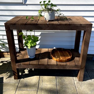 May include: A dark brown wooden table with two shelves. The table has a rustic look and is made of reclaimed wood. There are two potted plants on the top shelf and a wooden tray on the bottom shelf.