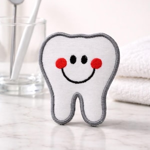 Peut inclure: Un jouet en peluche blanc en forme de dent avec un contour gris, des yeux noirs et des joues rouges. Un visage souriant est brodé sur le devant. Une brosse à dents, un miroir dentaire et des serviettes sont en arrière-plan.