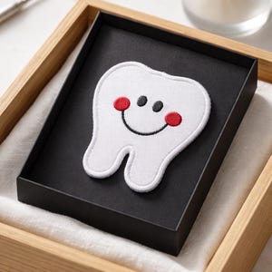 May include: A white, smiling tooth-shaped patch with red cheeks and a black smile and eyes, set in a black box within a wooden tray. The patch is an embroidered design, perfect for dental-themed crafts or gifts.