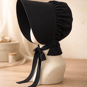 May include: A solid black bonnet featuring a wide, stiffened brim and a gathered crown. The headwear includes long, matching black fabric ties that are knotted under the chin, providing a secure fit. The design is simple, functional, and opaque.