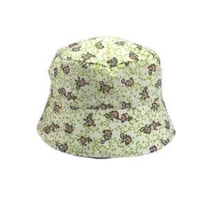May include: A light green bucket hat with a repeating pattern of cartoon monkeys and green vines. The hat is made of fabric and has a rounded crown and a downward-sloping brim. The hat is designed to be worn on the head.