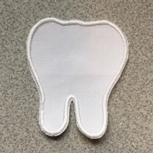 May include: A white fabric tooth-shaped patch with a white embroidered border. The patch is a bright white colour. The background is a speckled grey surface.