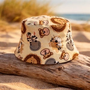 May include: A beige bucket hat featuring a repeating pattern of cartoon animals, including lions, monkeys, zebras, and tigers. The hat is made of fabric and sits on a piece of driftwood on a sandy beach.