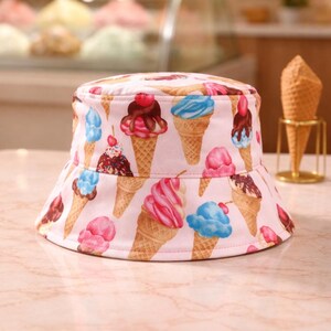 May include: A pink bucket hat featuring a repeating pattern of ice cream cones with various flavors. The ice cream cones have chocolate, cherry, and blue and pink ice cream. The hat is on a marble surface.